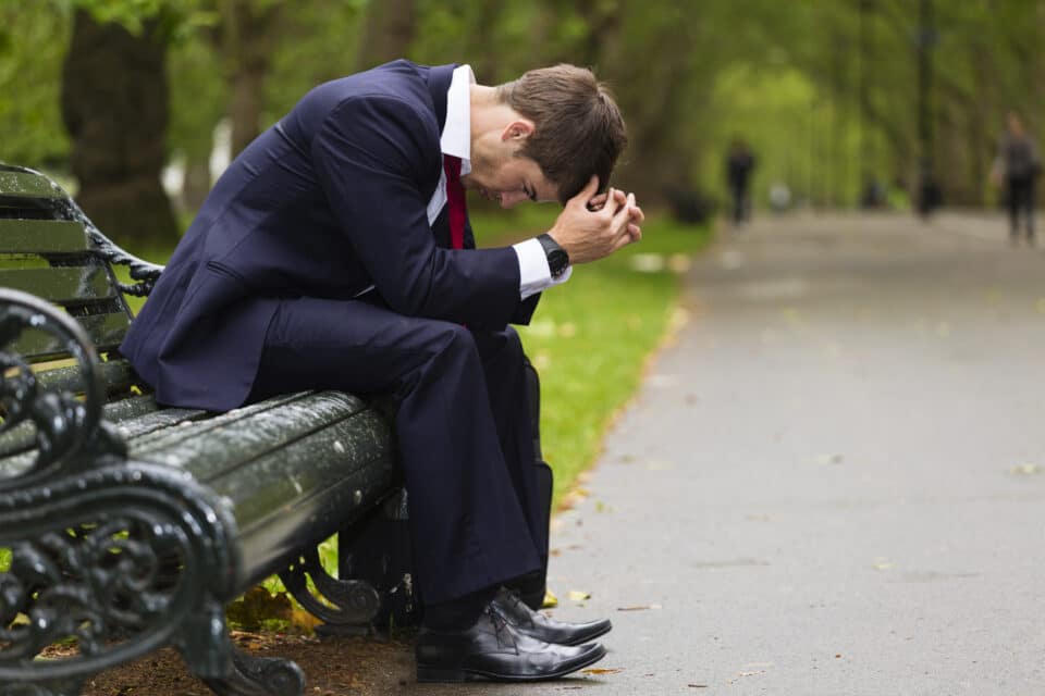Man in a suit sitting on a park bench with his head bowed and hands clasped, appearing stressed or upset.