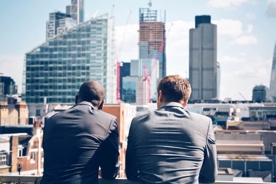 Two businessmen in suits standing on a balcony, looking out over a city skyline with modern high-rise buildings.