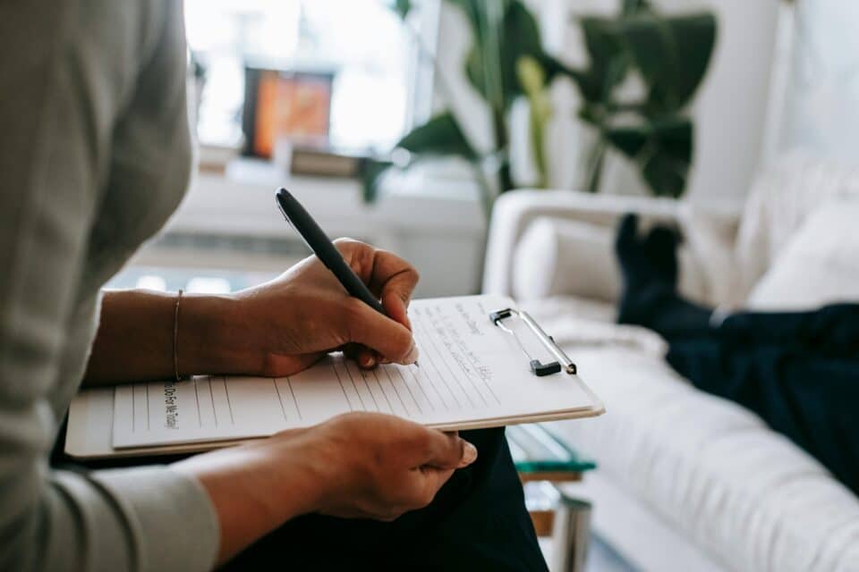 Person writing notes on a clipboard during a session, with another person lying on a sofa in the background.