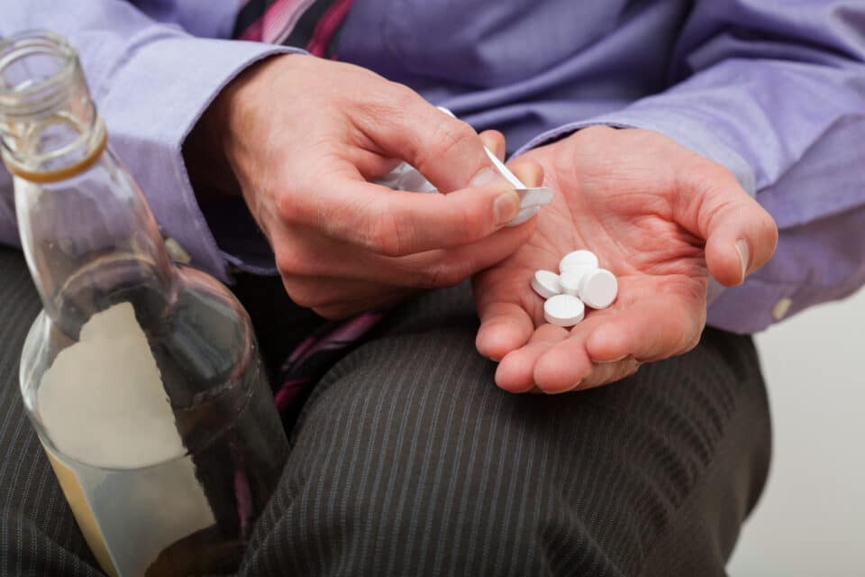 "Person holding several white pills in one hand and an alcohol bottle in the other, suggesting substance misuse.
