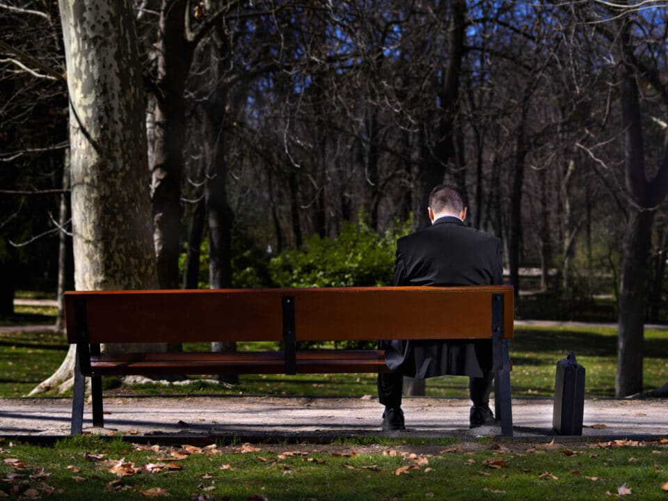 Man in a suit sitting alone on a park bench, with a briefcase beside him.