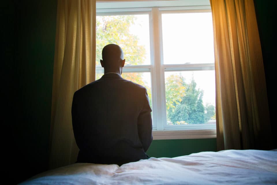 A person in a suit sits on the edge of a bed, facing a bright window with trees outside, creating a reflective and contemplative mood.