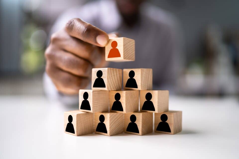 A person stacking wooden blocks with human icons, symbolising leadership, teamwork, and organisational structure.
