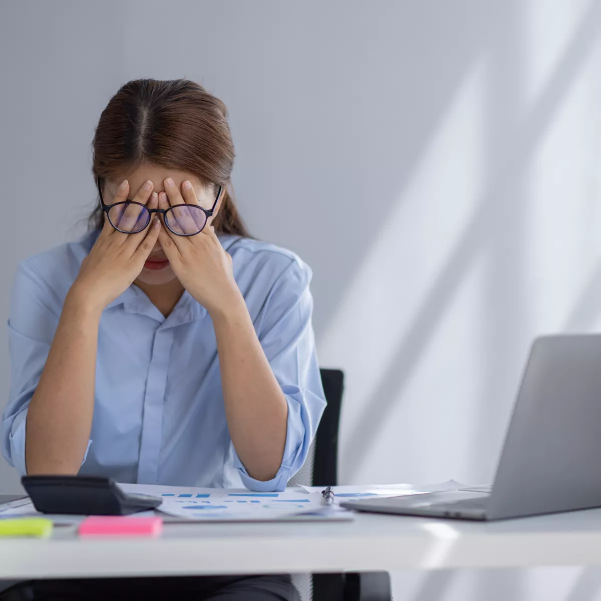 A woman in a blue shirt sits at a desk with her head in her hands, looking stressed or overwhelmed. She is surrounded by a laptop, documents, and office supplies, with sunlight casting shadows on the wall behind her.