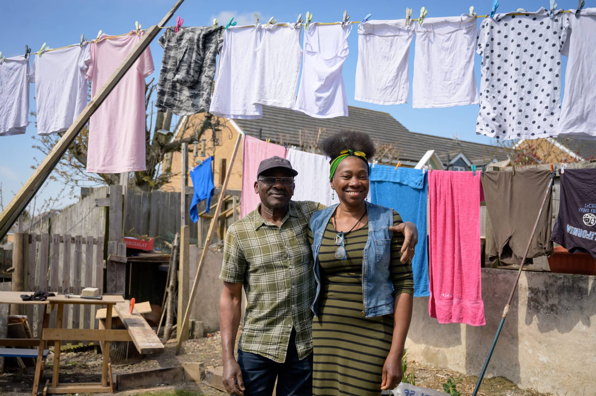 Smiling man and woman standing together in a backyard with laundry hanging on clotheslines behind them on a sunny day.
