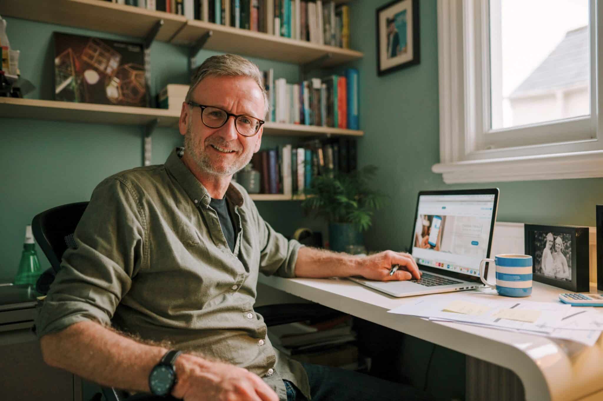 Older man wearing glasses, smiling while working on a laptop at a desk in a home office with bookshelves and personal photos in the background.