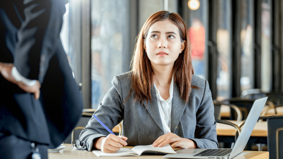 A woman in a grey business suit sits at a desk with a notebook and laptop, looking up at a standing colleague with a concerned or uncomfortable expression. The image appears to show a tense workplace interaction in an office setting.
