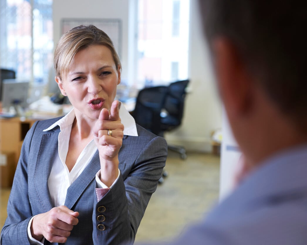 Businesswoman in a grey suit pointing and speaking sternly at a colleague in an office, appearing angry or confrontational.