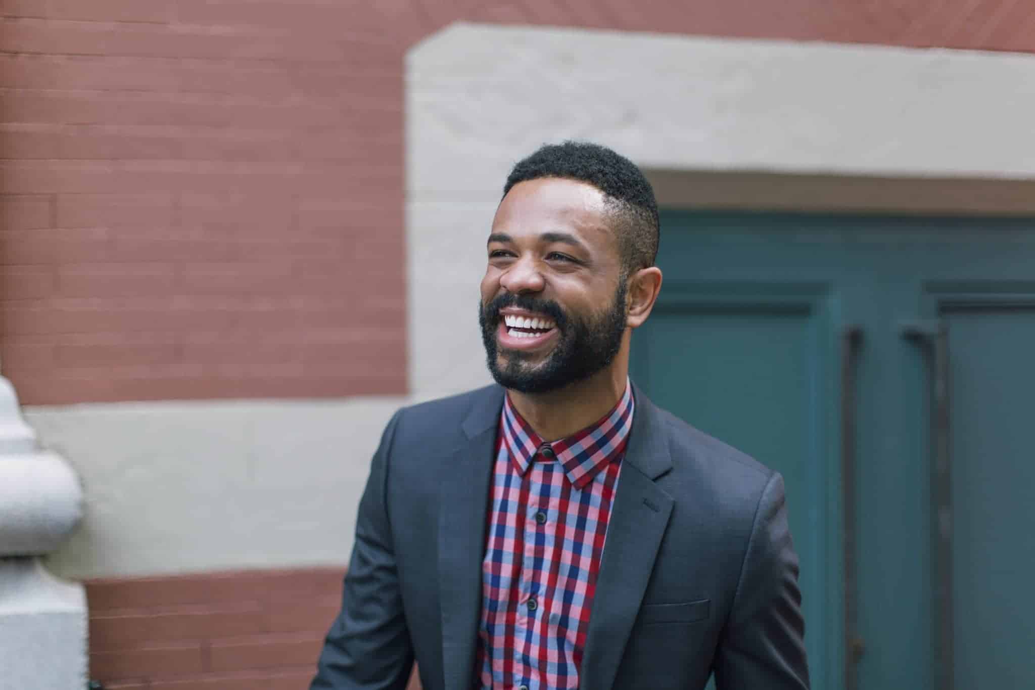 Smiling man wearing a dark suit jacket and red-and-blue checkered shirt standing outdoors against a brick wall.