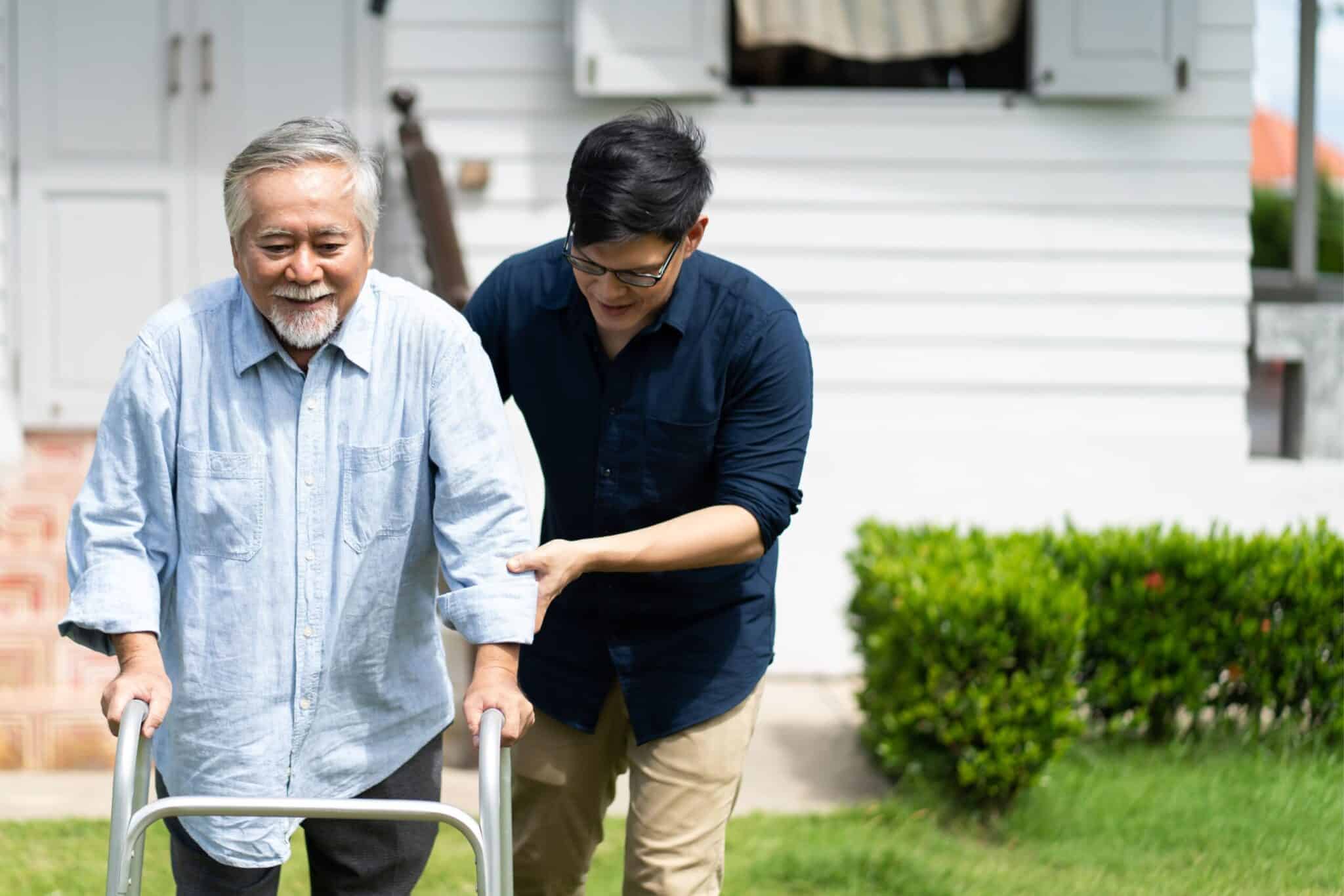 Young man supporting an elderly man using a walking frame outdoors.