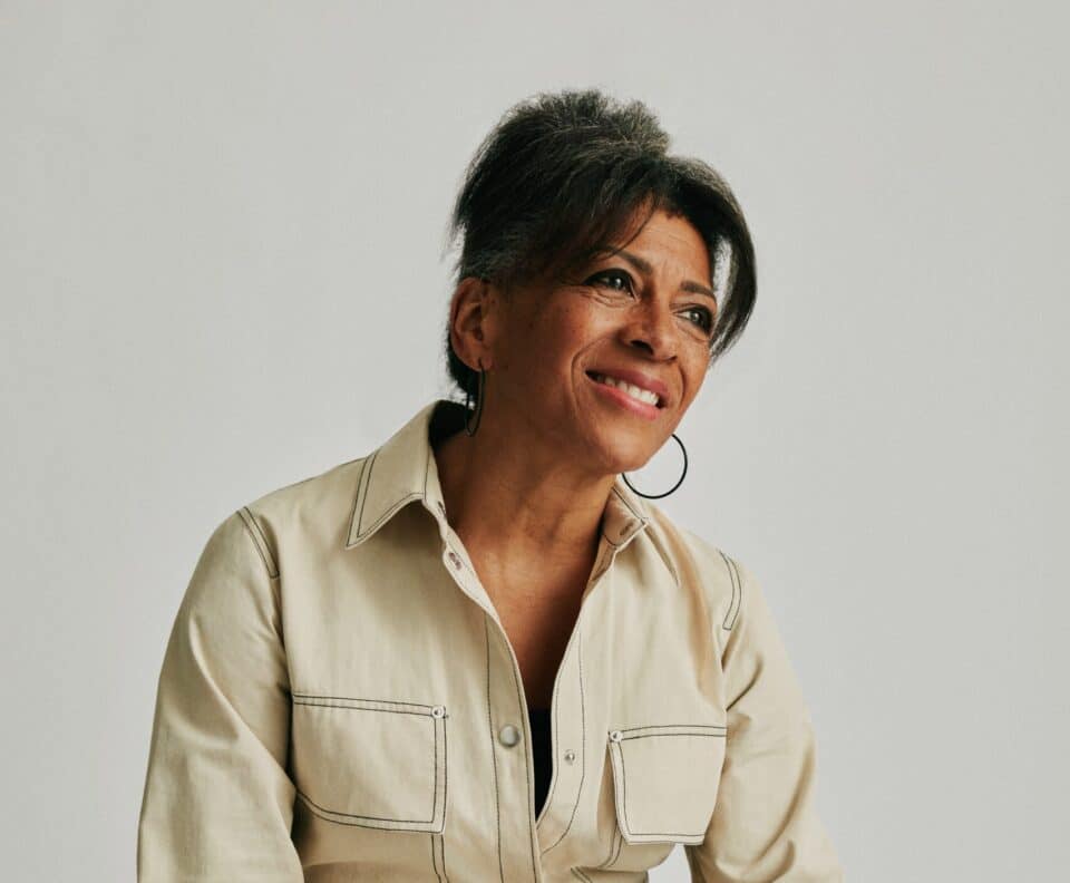 An older woman with dark hair tied back, wearing a light beige jumpsuit with dark stitching, sits on a wooden stool smiling and looking slightly upward. She has hoop earrings and several rings on her fingers.