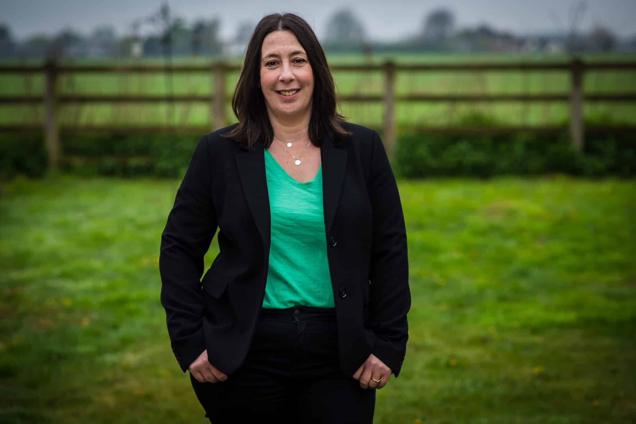 Woman in green top and black suit smiling, stood in front of a field and a fence - Claudia De Silva