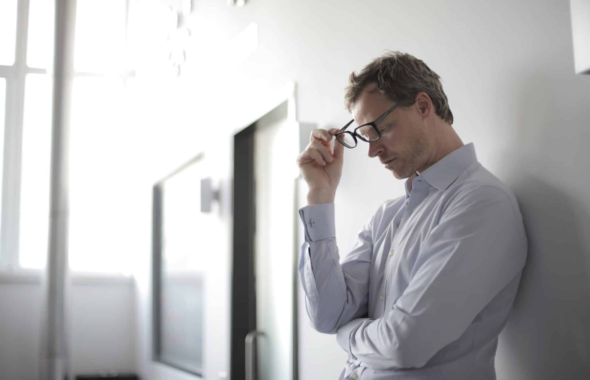 Man with glasses wearing a shirt leaning against a wall in an office looking worried and tired