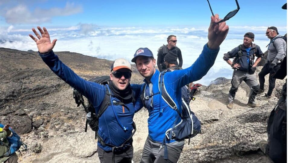 Two men in blue tops celebrating at the top of a mountain