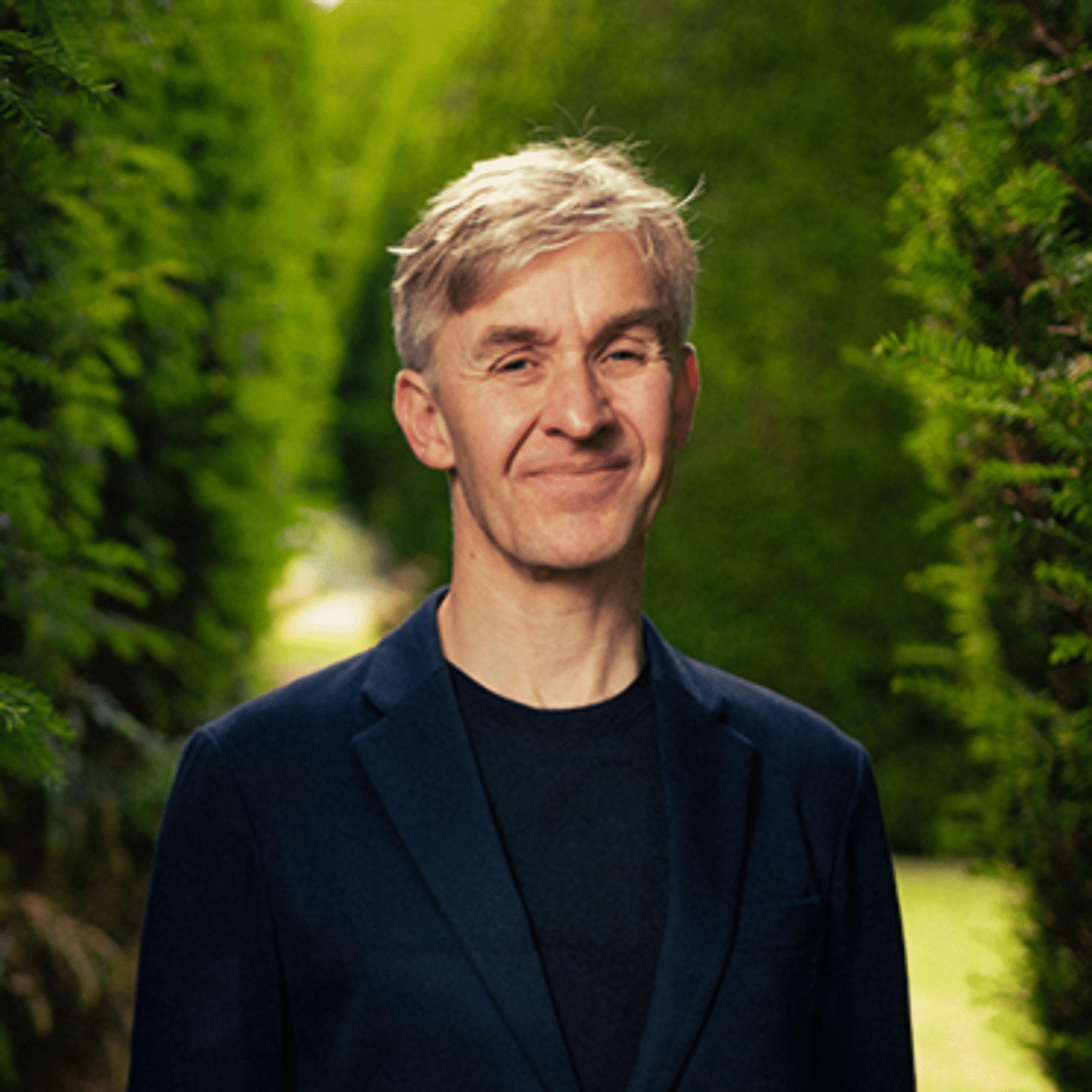 Man with short blond hair smiling stood in front of some trees