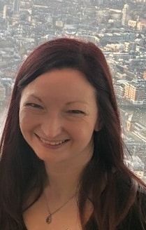Woman with long dark hair smiling and stood in front of window looking out over a city - Tanya Dolan