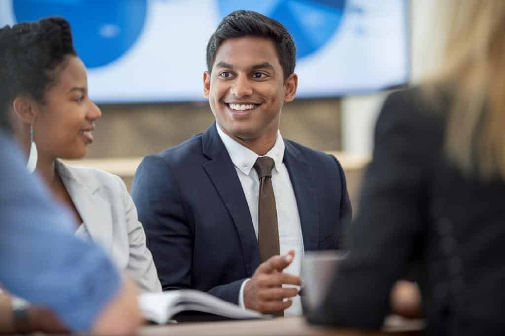 Young man in suit say with other people at a table looking happy.