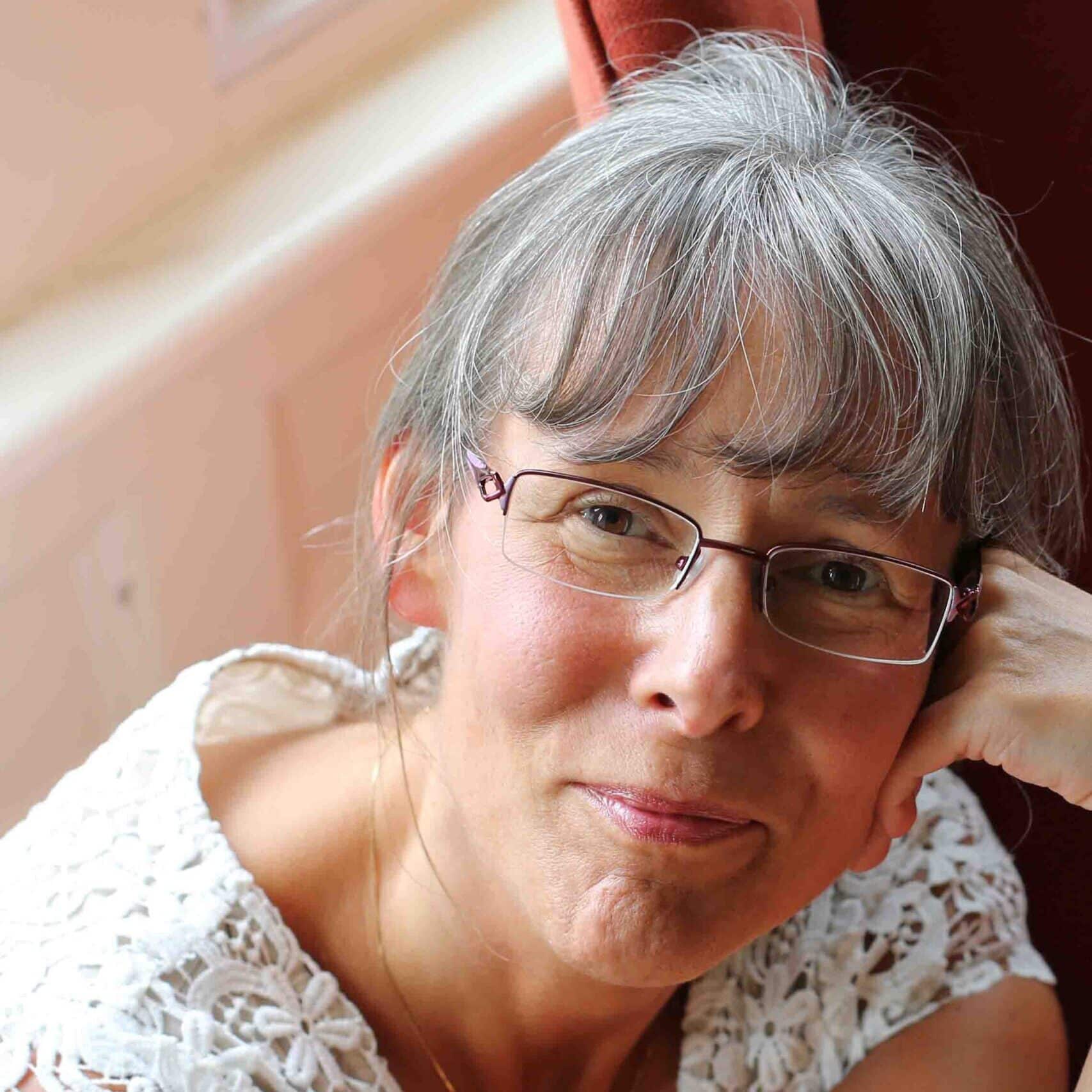 Woman with gray hair and glasses, wearing a white lace top, smiling gently while resting her head on her hand.
