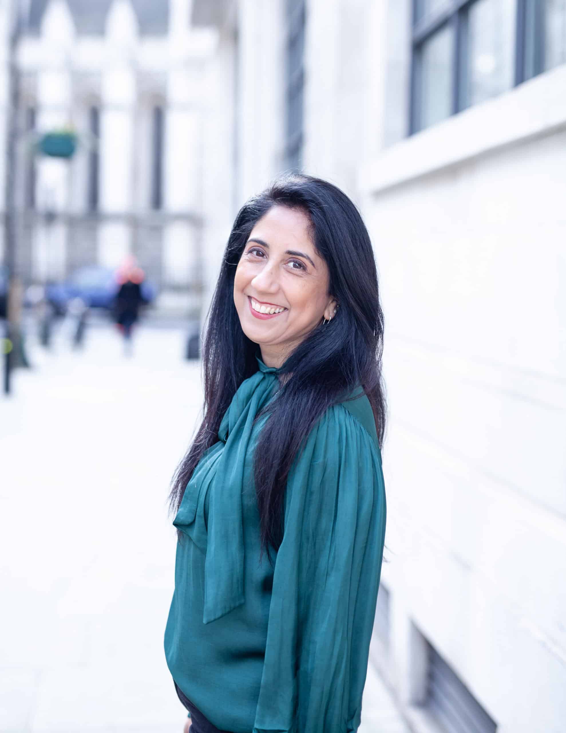 Smiling woman with long dark hair wearing a green blouse, standing outdoors by a white stone building and looking towards the camera.