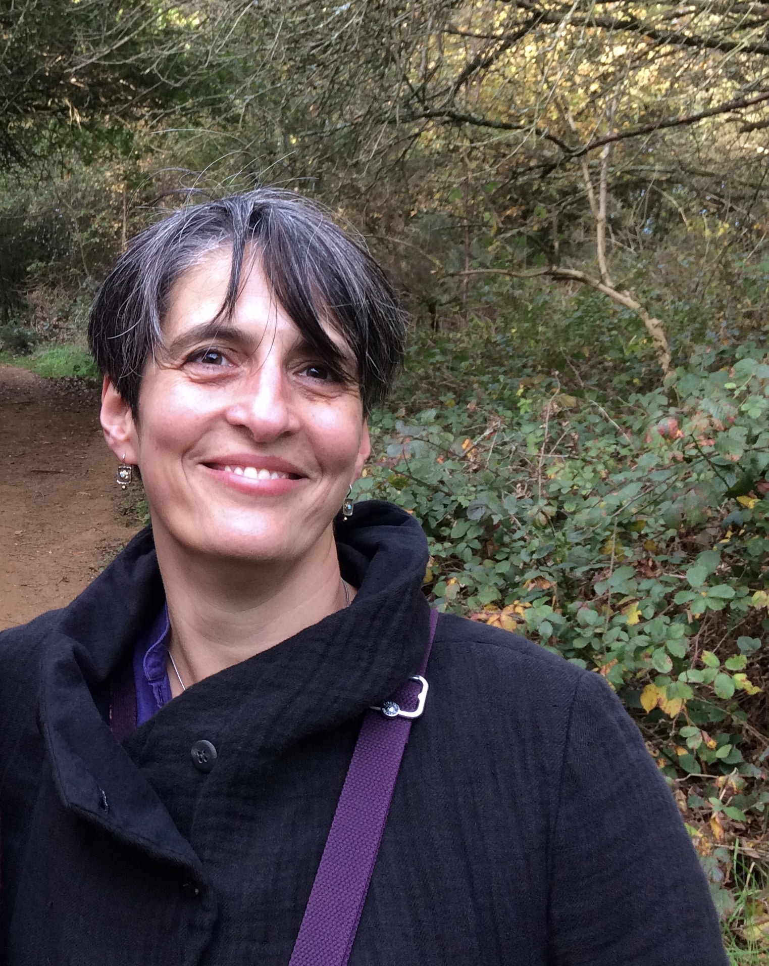 Smiling woman with short dark and grey hair wearing a black coat, standing on a woodland path.