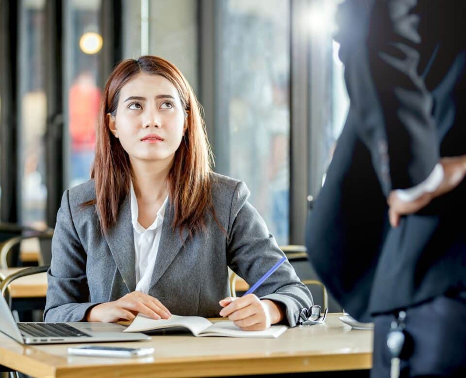 A woman in a gray suit sitting at a desk with a notebook and laptop, looking up with a worried or uneasy expression at a person standing nearby, suggesting a tense or uncomfortable interaction.