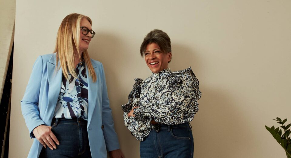 Two older women smiling and standing side by side against a plain beige wall. The woman on the left has long blonde hair, wears glasses, a light blue blazer, a patterned blouse, and dark jeans. The woman on the right has short gray hair, wears a floral ruffled blouse and dark jeans. Both appear relaxed and cheerful.