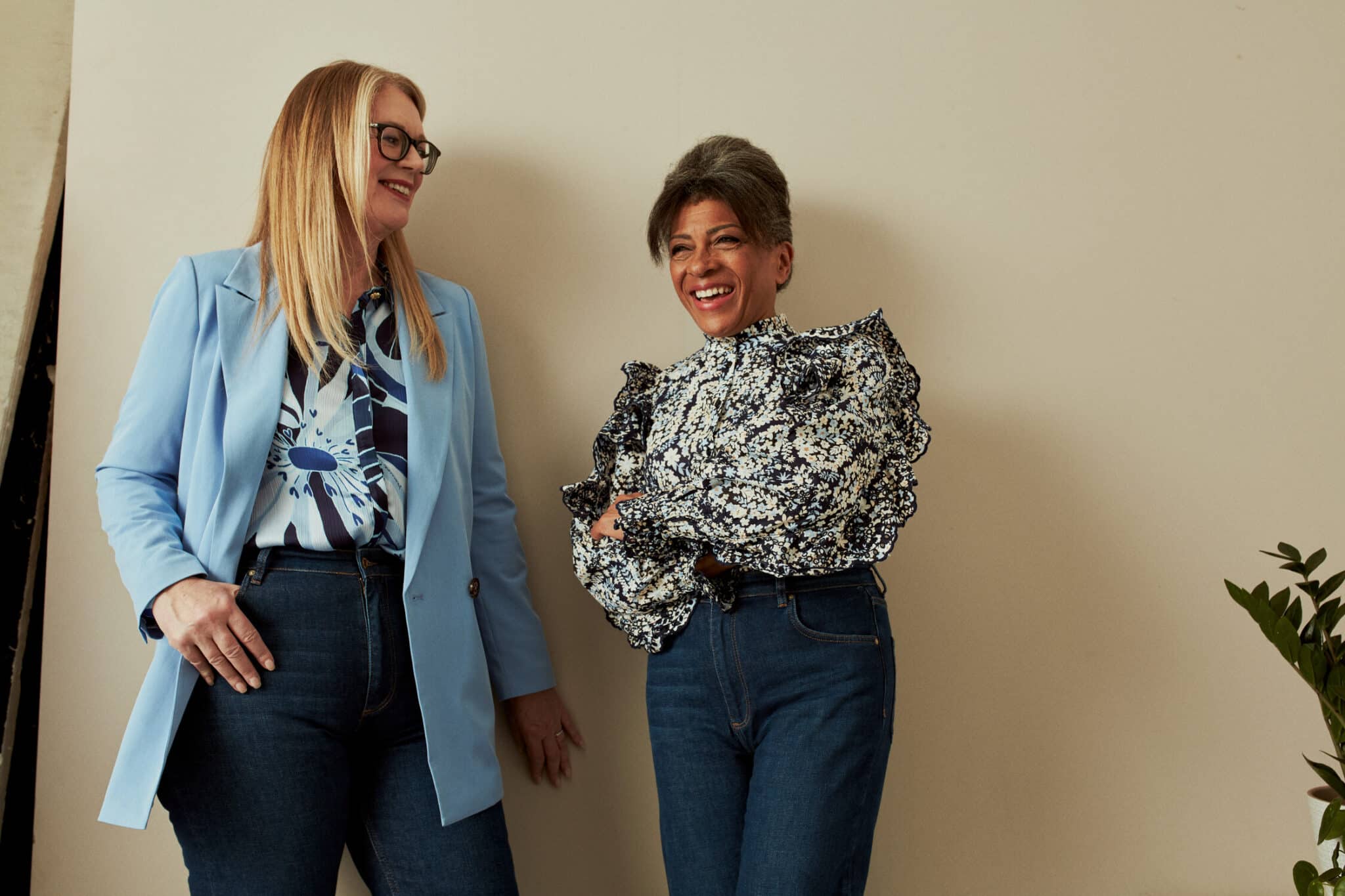 Two older women smiling and standing side by side against a plain beige wall. The woman on the left has long blonde hair, wears glasses, a light blue blazer, a patterned blouse, and dark jeans. The woman on the right has short gray hair, wears a floral ruffled blouse and dark jeans. Both appear relaxed and cheerful.