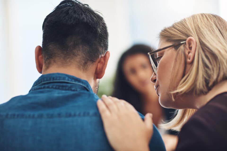 Woman with glasses gently resting her hand on a man’s shoulder, offering comfort and support, with another person blurred in the background.