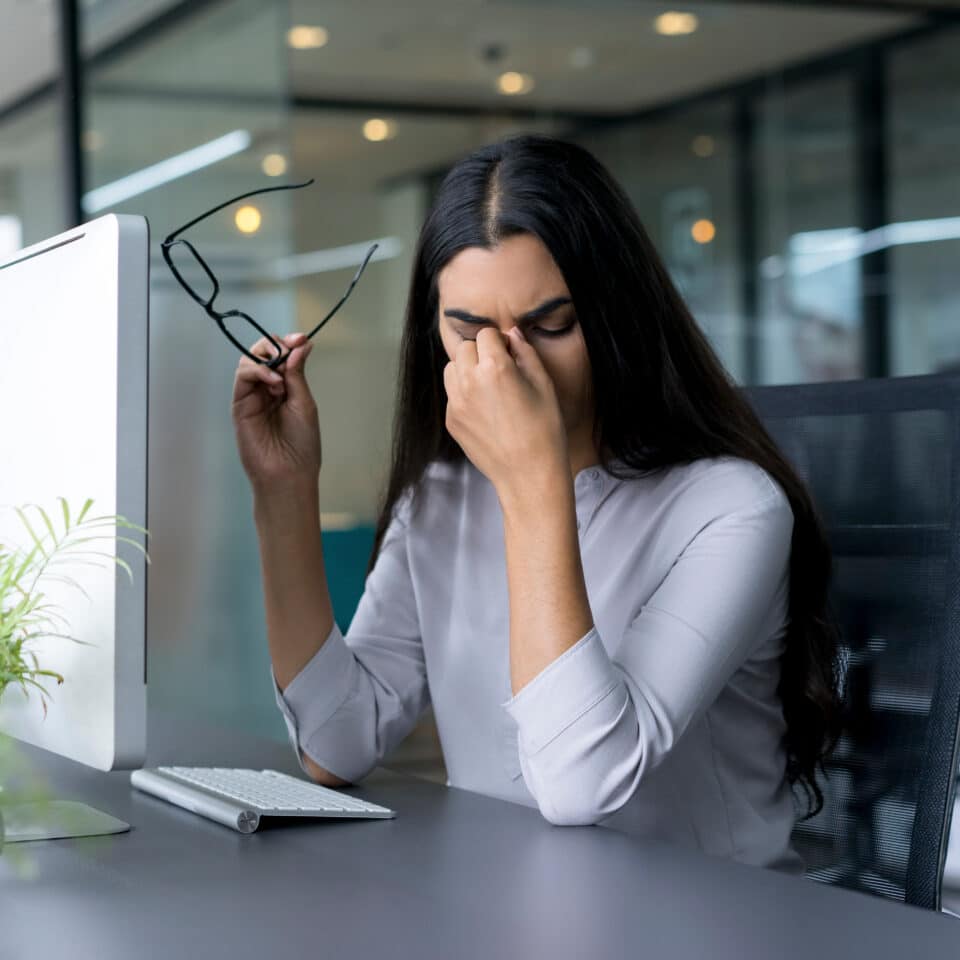 A woman sits at a desk in an office, holding her glasses in one hand and pinching the bridge of her nose with the other, looking stressed or fatigued in front of her computer.