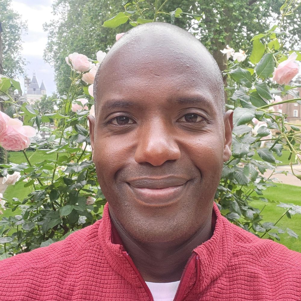 Man smiling at the camera, stood outside in front of a pink rose bush