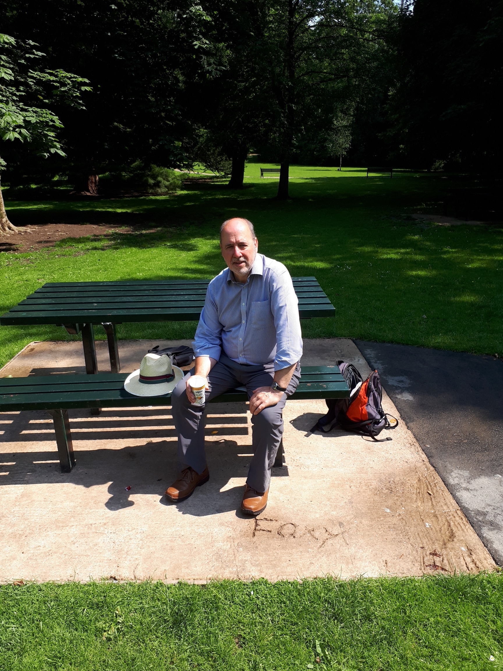 A man sitting on a green park bench holding a coffee cup on a sunny day, with a hat beside him and a backpack on the ground, surrounded by grass and trees.