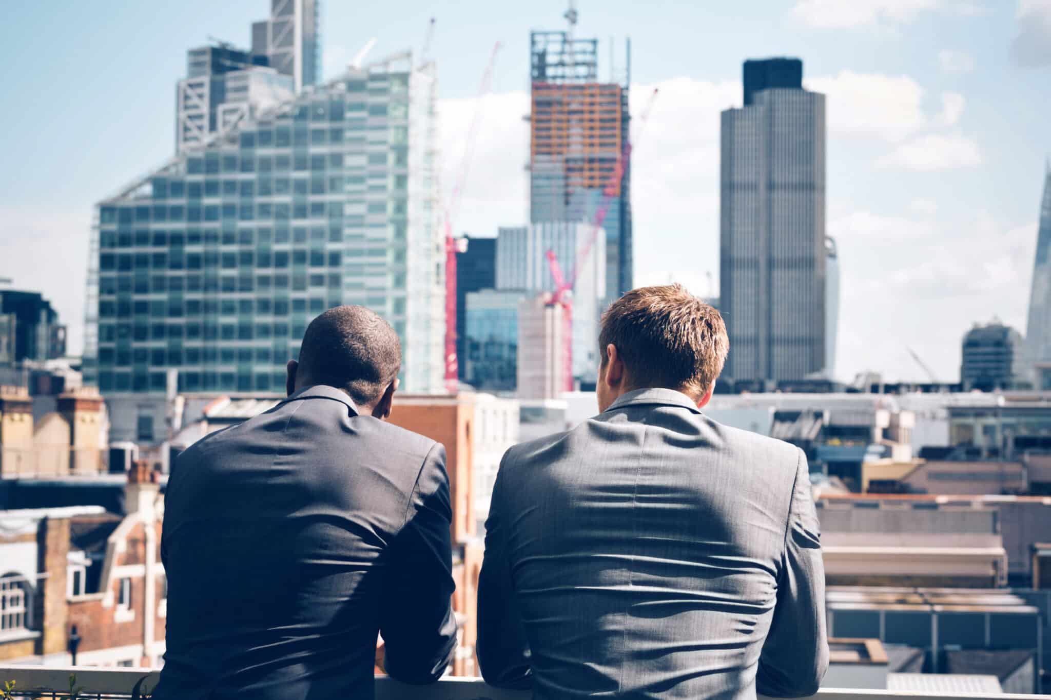 Two businessmen in suits standing on a balcony, looking out over a city skyline with modern high-rise buildings.