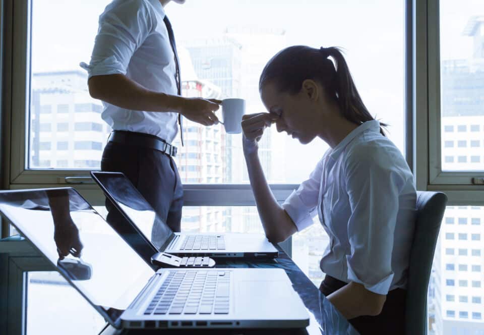 Stressed businesswoman sitting at a desk with two laptops, holding her head in frustration, while a colleague stands nearby holding a coffee cup.