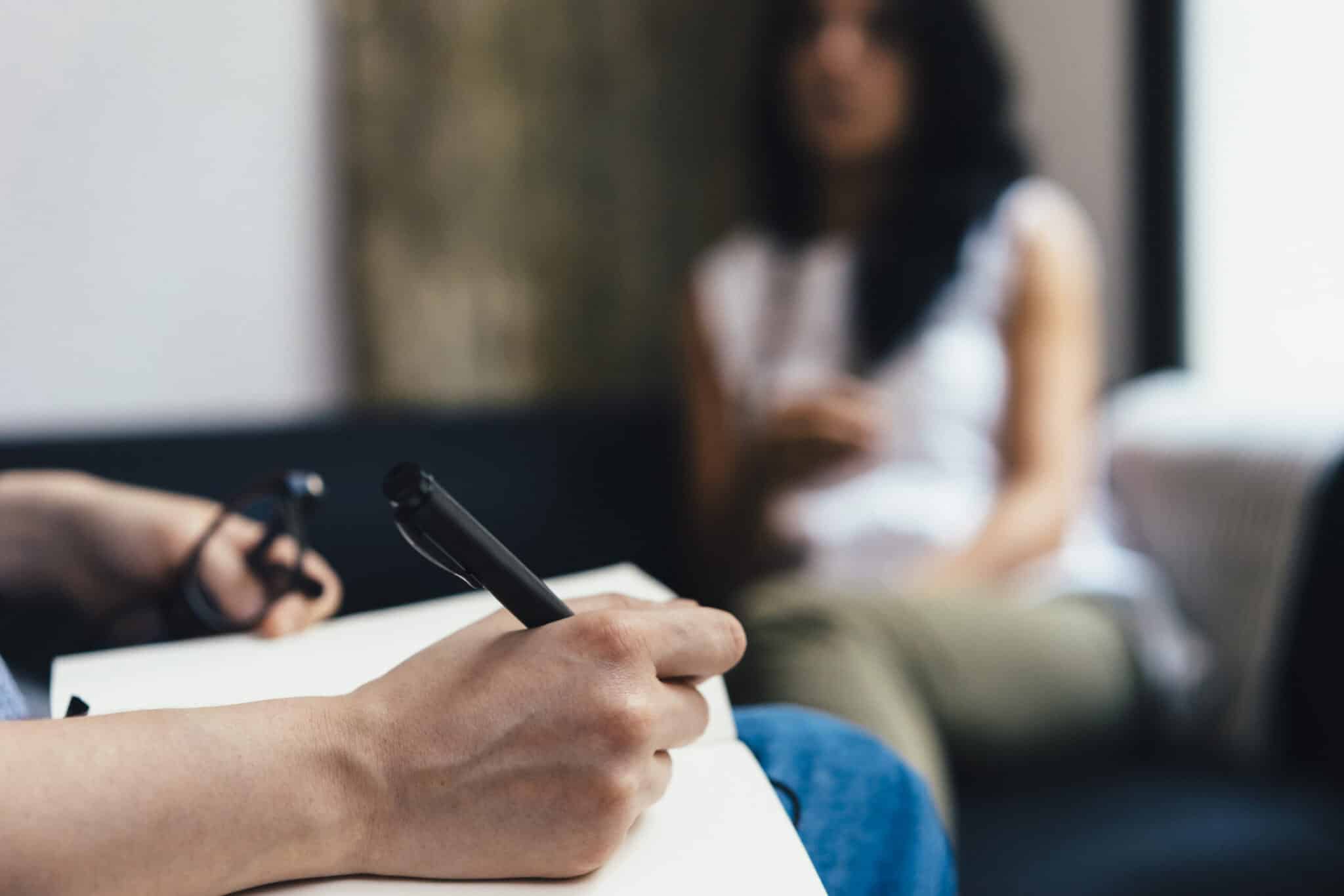 Close-up of a person writing notes during a conversation, with another person sitting blurred in the background, suggesting a counselling or support session.