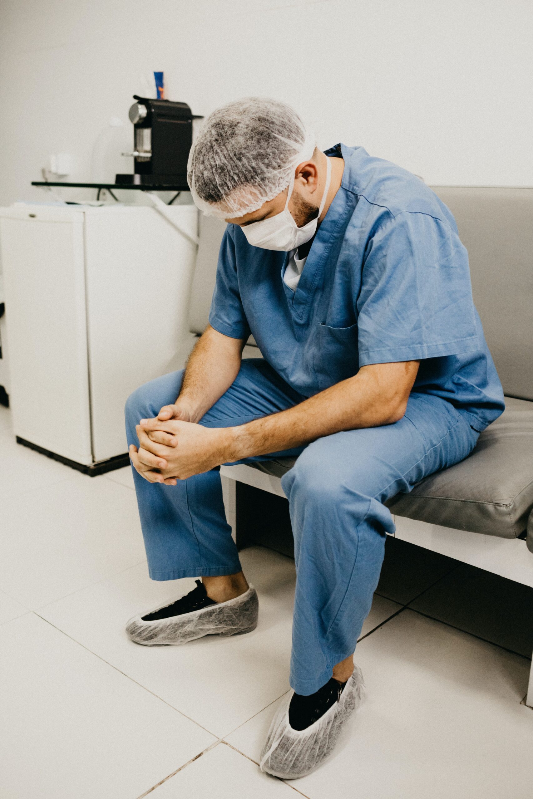 Tired healthcare worker in scrubs and a face mask sitting on a bench with head bowed and hands clasped.