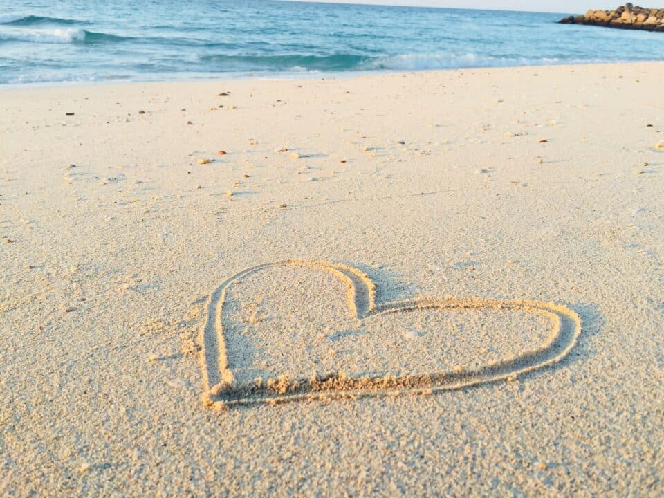 A heart shape drawn in the sand on a beach, with gentle ocean waves in the background.