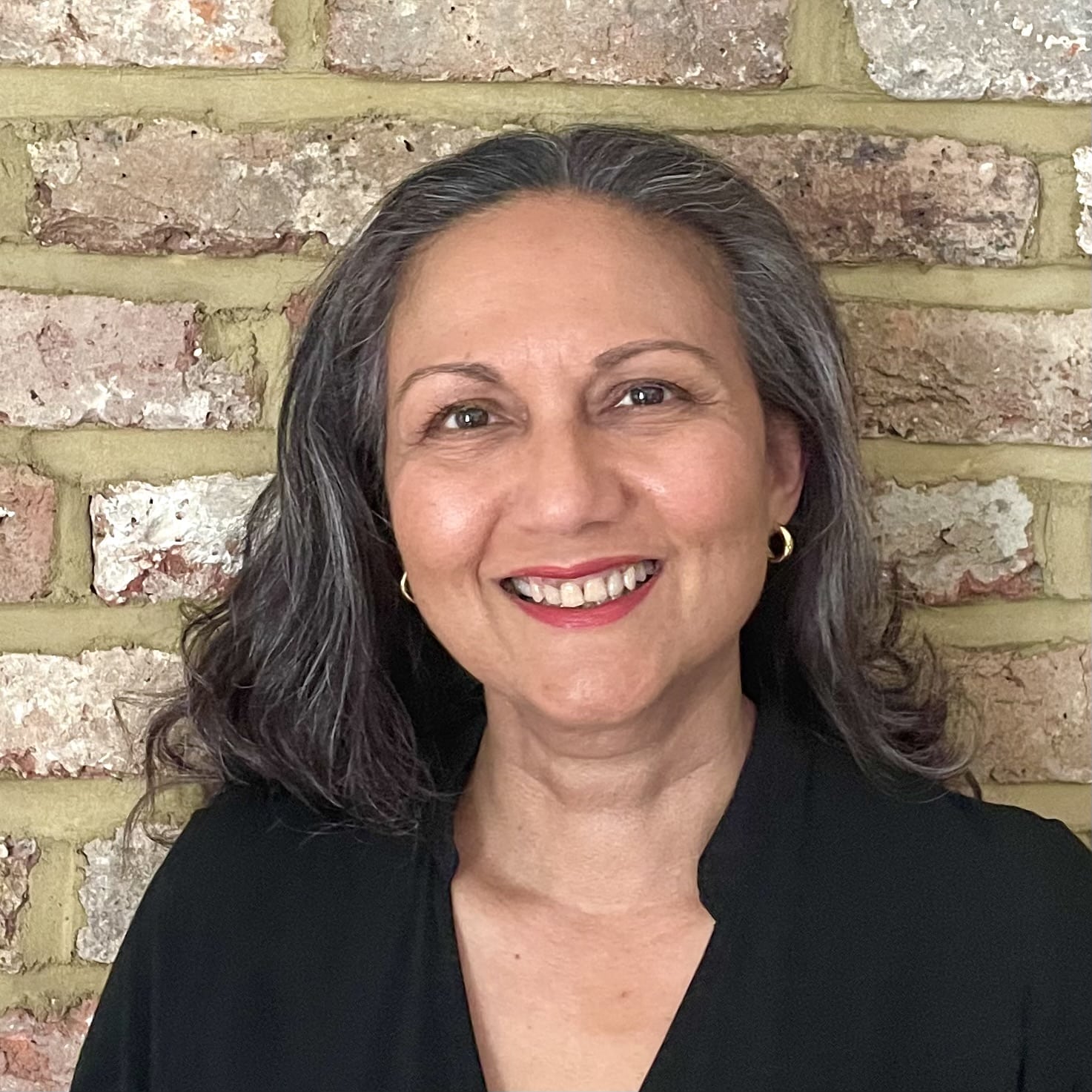 Smiling woman with shoulder-length dark hair and grey streaks, wearing a black top and gold hoop earrings, standing in front of a brick wall.