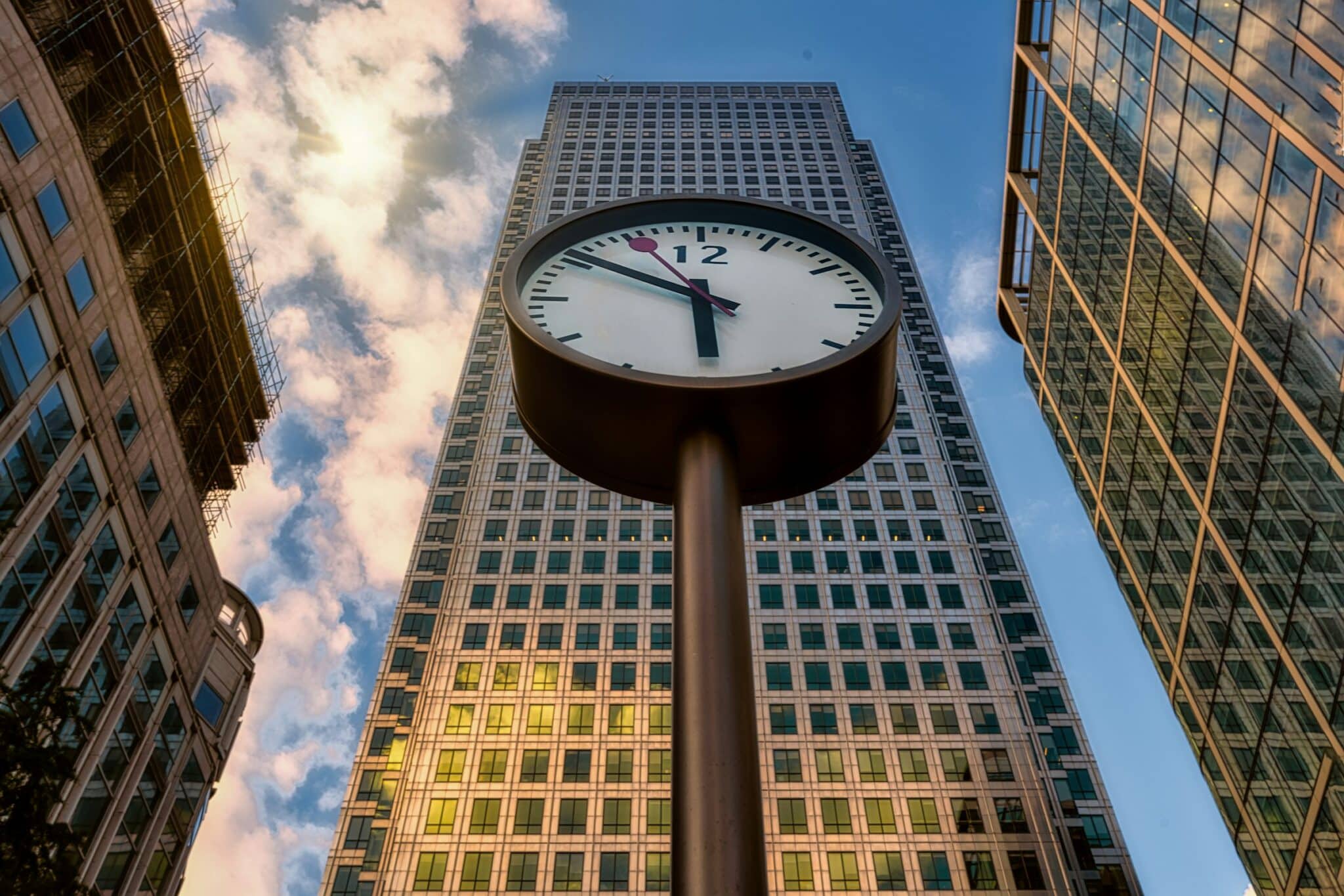 Large outdoor clock in a city financial district, viewed from below with tall glass office buildings and a partly cloudy sky in the background.