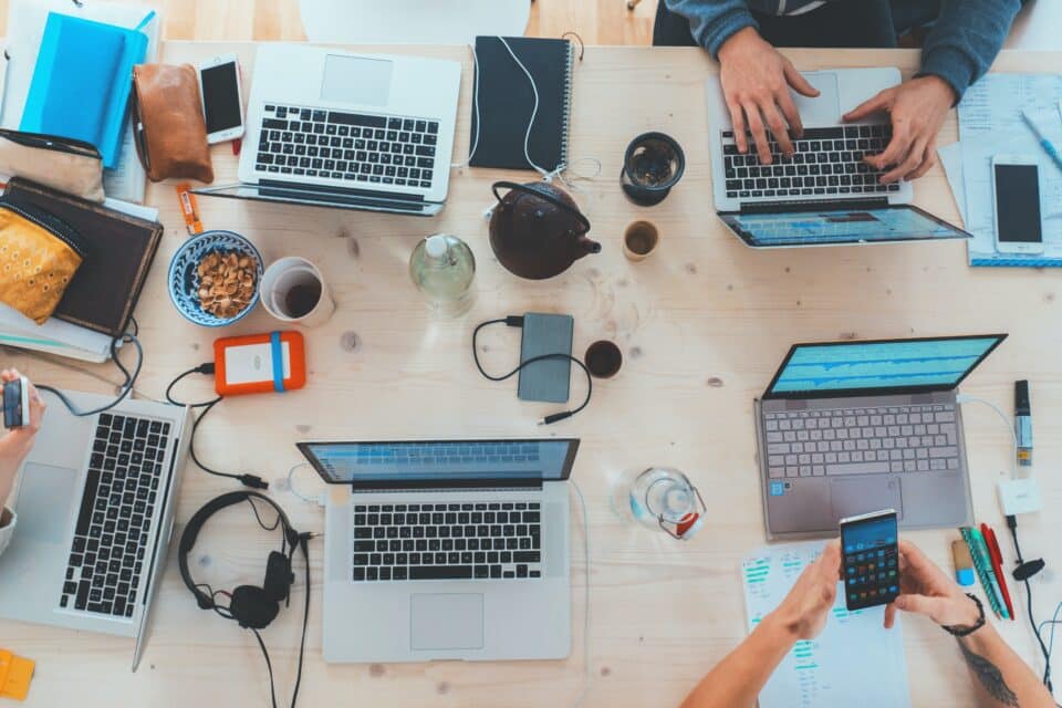 Overhead view of a busy shared workspace with several laptops, smartphones, notebooks, headphones, and drinks; hands typing and holding a phone.