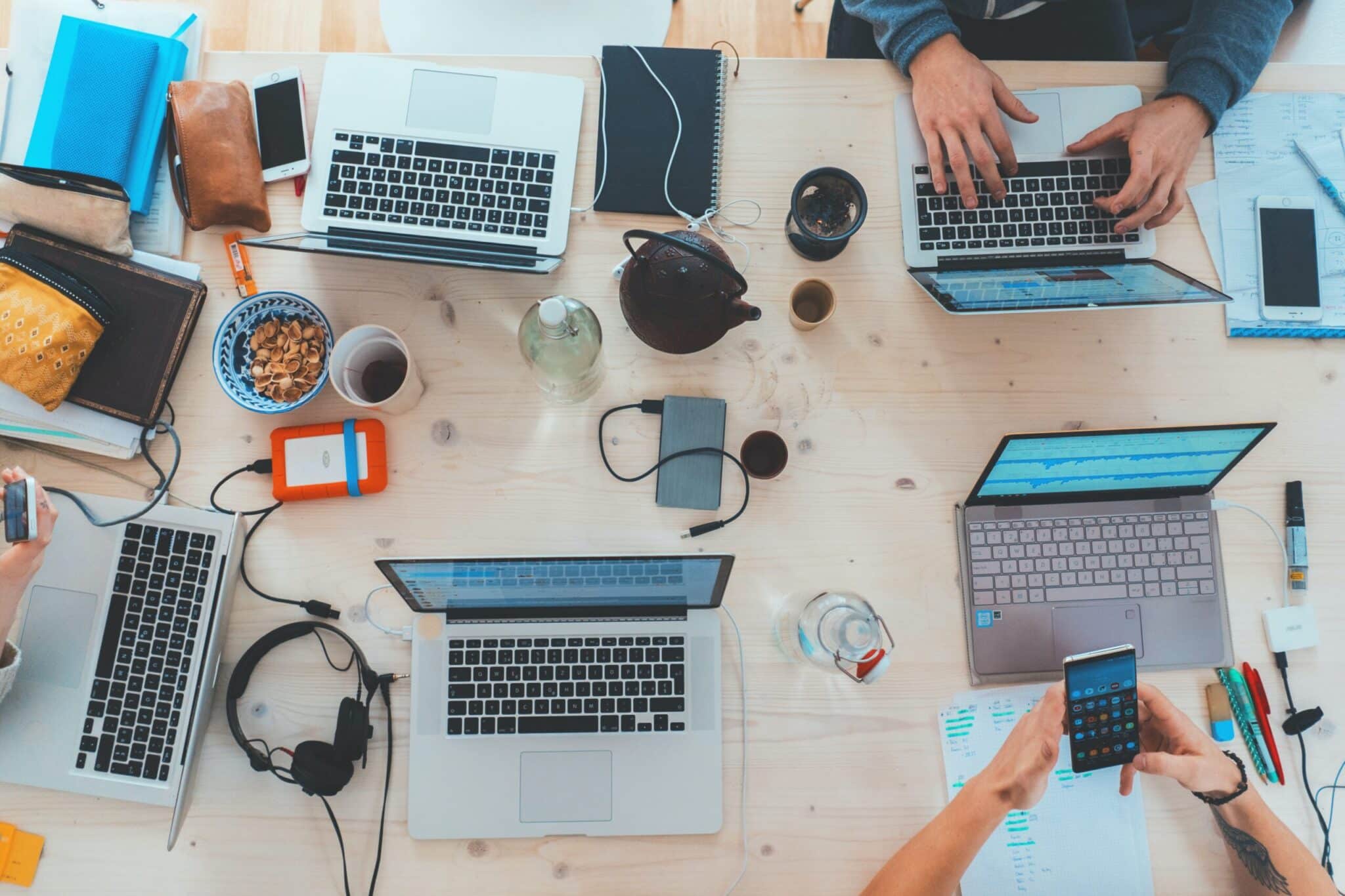 Overhead view of a busy shared workspace with several laptops, smartphones, notebooks, headphones, and drinks; hands typing and holding a phone.