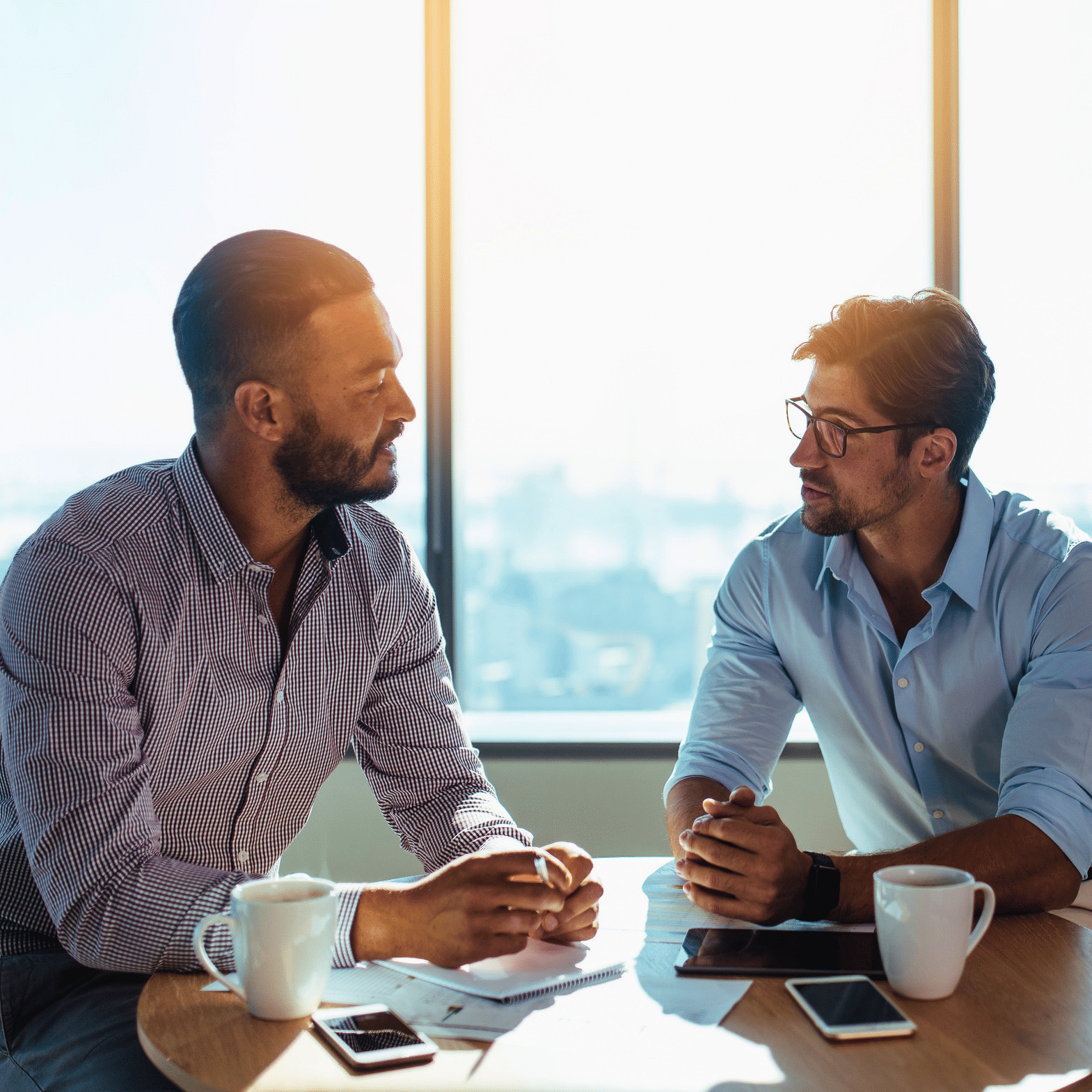 Two men sitting at a round table in a bright office, having a serious conversation over coffee, with notebooks and smartphones on the table.