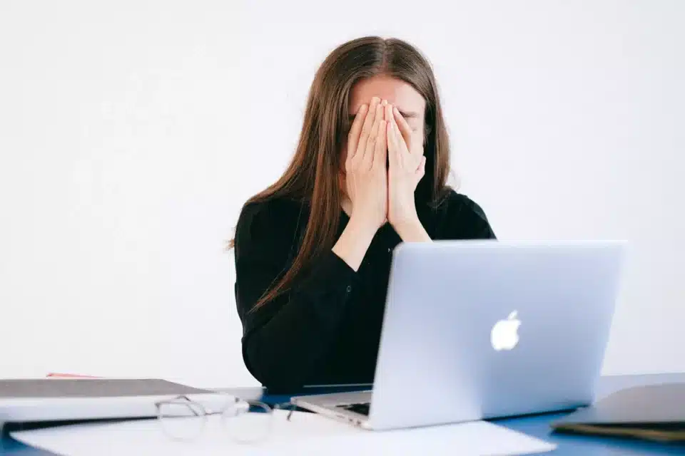 Young woman sat at desk in front of laptop with her face in her hands.