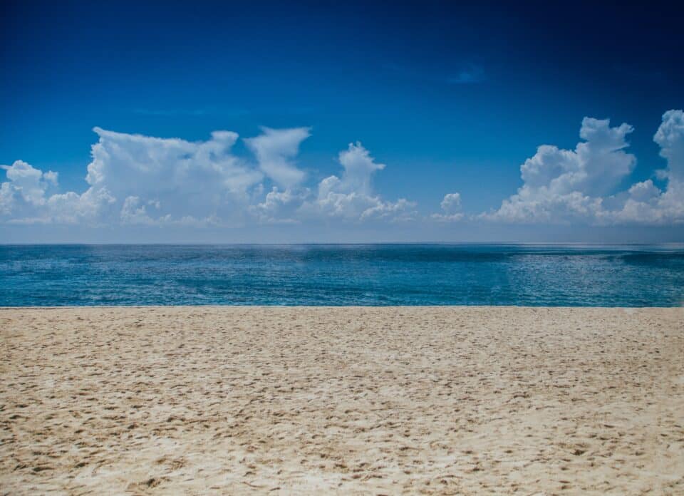 A peaceful beach scene with golden sand in the foreground, calm blue ocean water stretching to the horizon, and fluffy white clouds scattered across a deep blue sky.