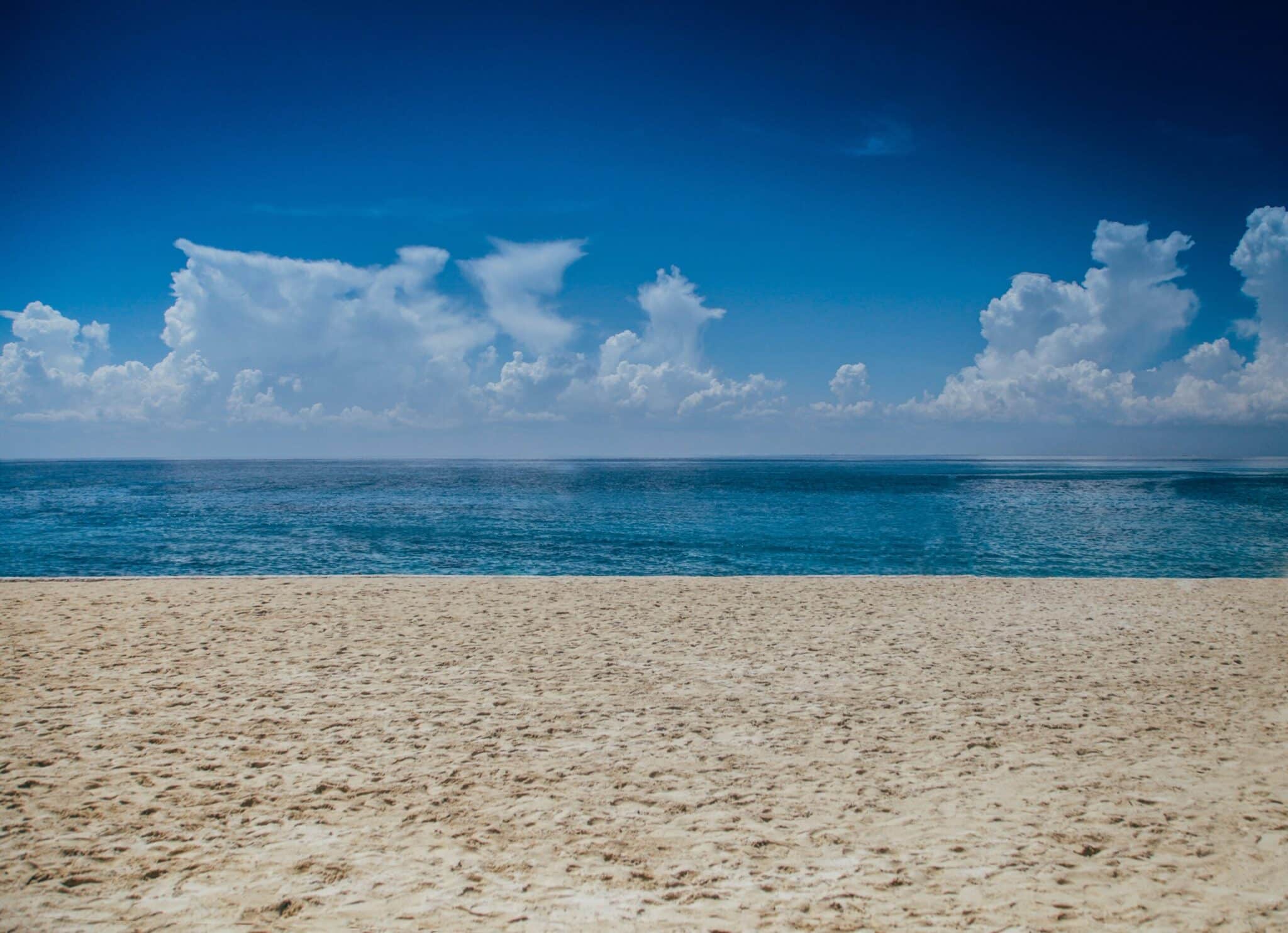 A peaceful beach scene with golden sand in the foreground, calm blue ocean water stretching to the horizon, and fluffy white clouds scattered across a deep blue sky.