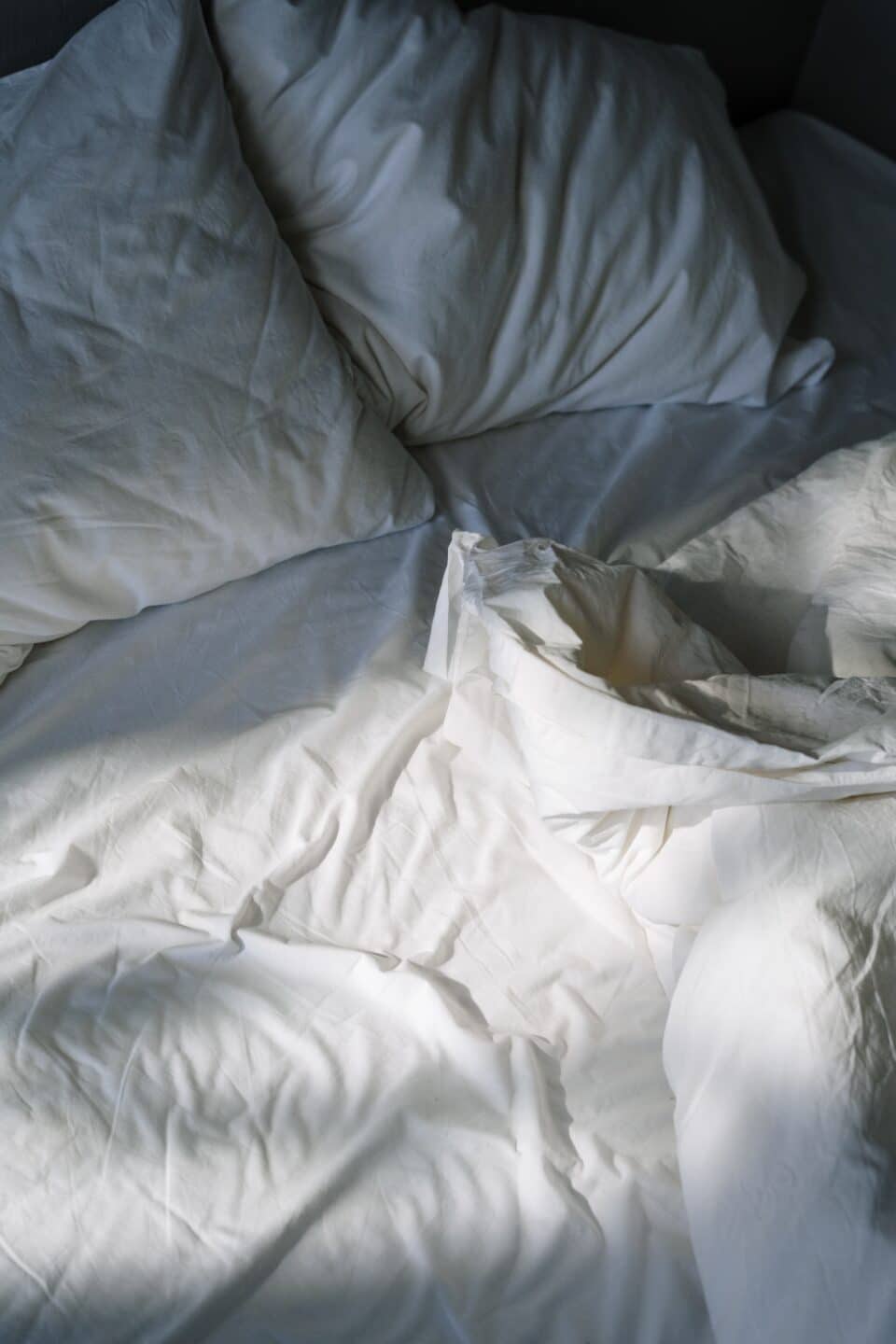 A messy unmade bed with white pillows and sheets, softly lit by natural light coming through a nearby window.