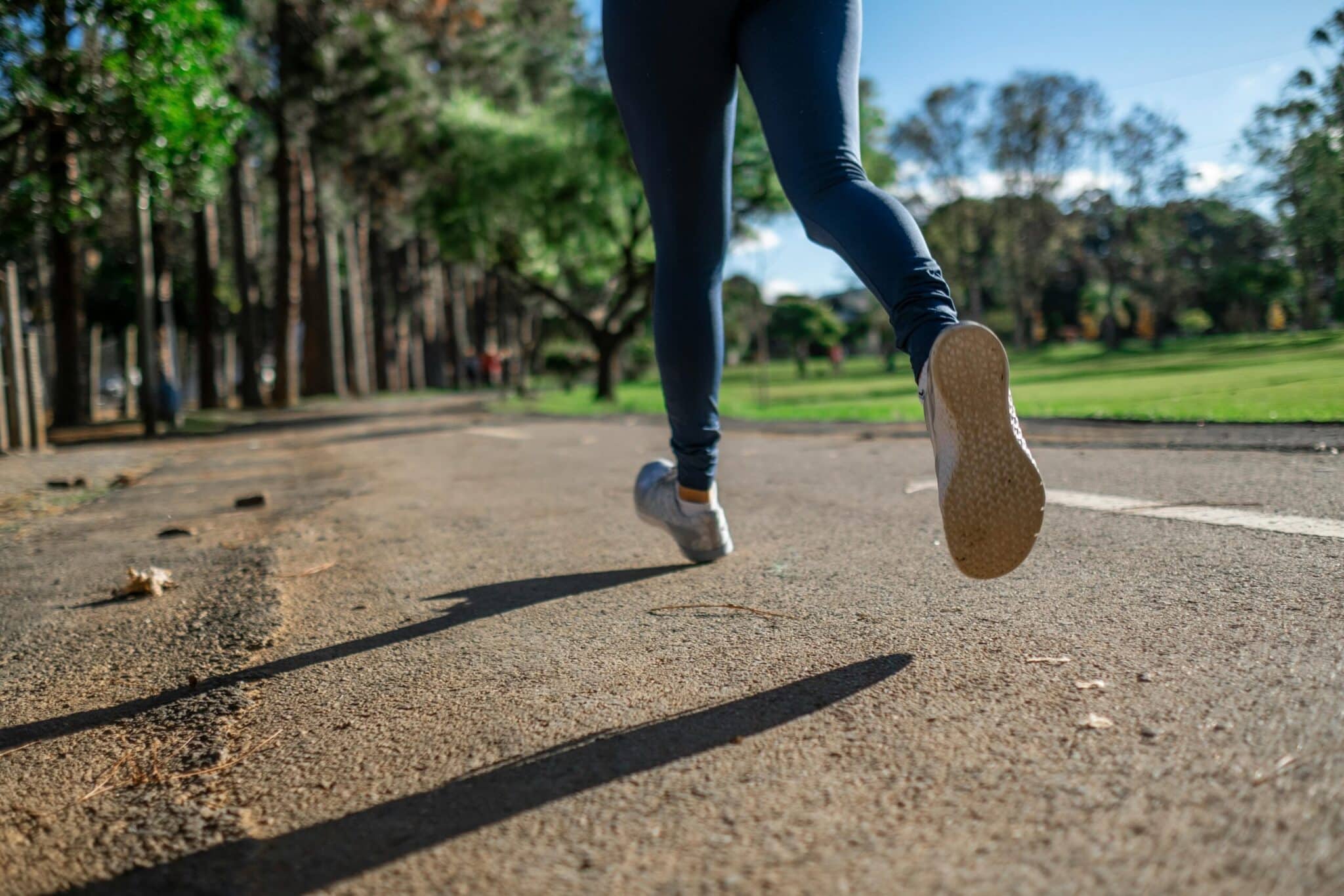 Person jogging outdoors on a sunny day along a tree-lined path, focusing on their legs and running shoes.