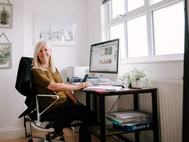 Smiling woman sitting at a desk in a bright home office, working on a computer with papers and plants around her.
