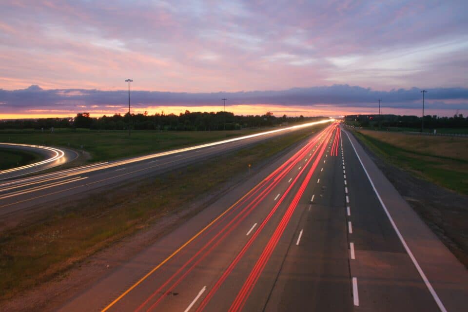 Long-exposure photo of a highway at sunset, showing streaks of red and white lights from passing vehicles under a colorful sky.