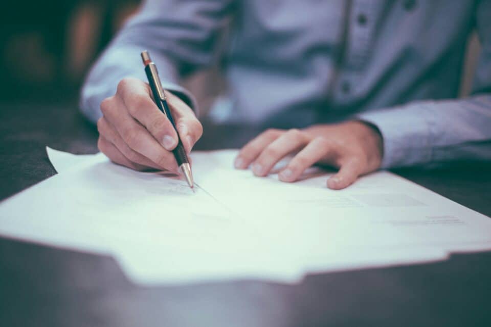 A person in a blue shirt writing on a stack of papers with a pen at a desk, focusing on work or signing documents.