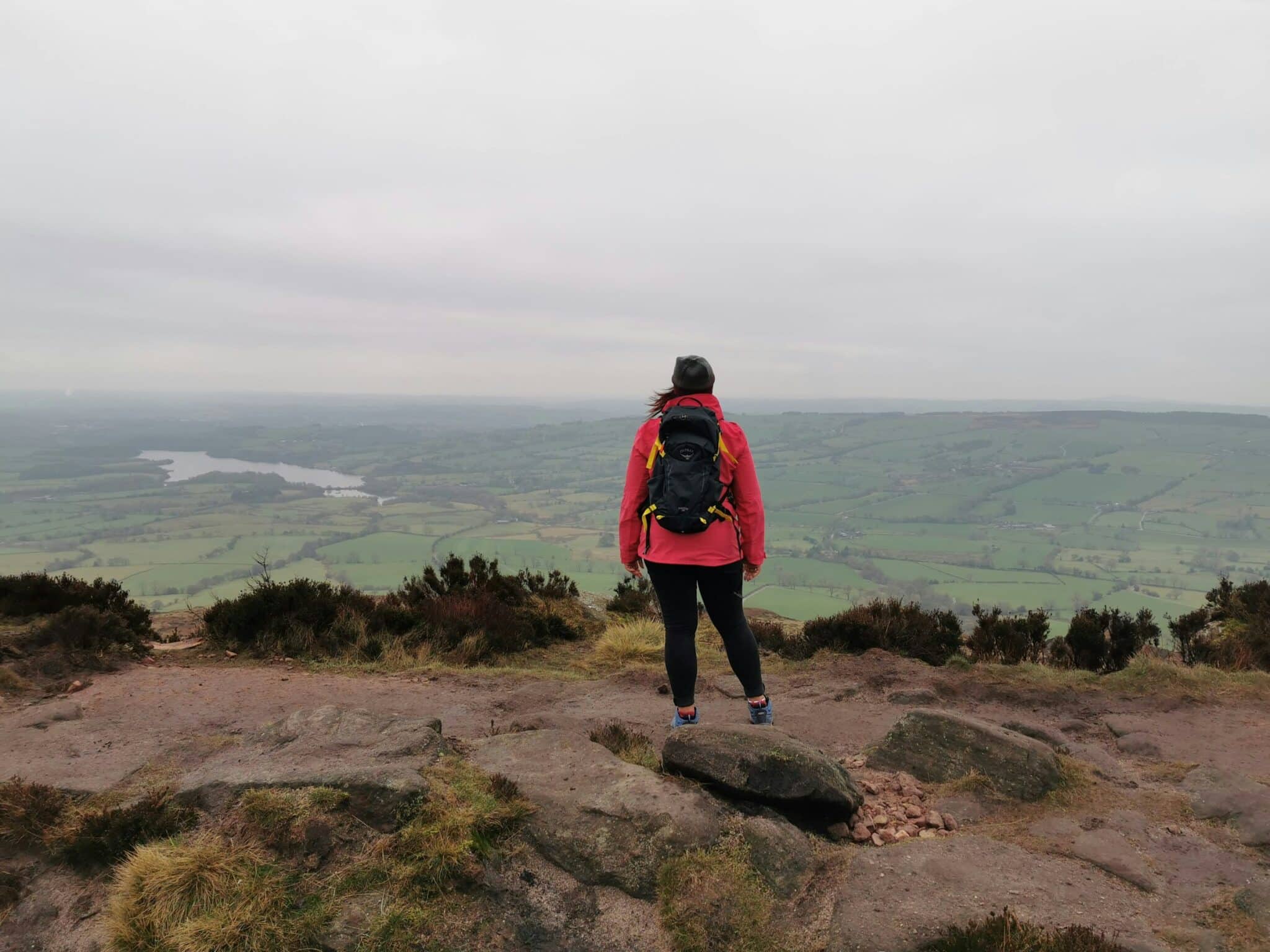 Woman with orange jacket and rucksack on top of hill looking out out over a valley with a lake in the far background